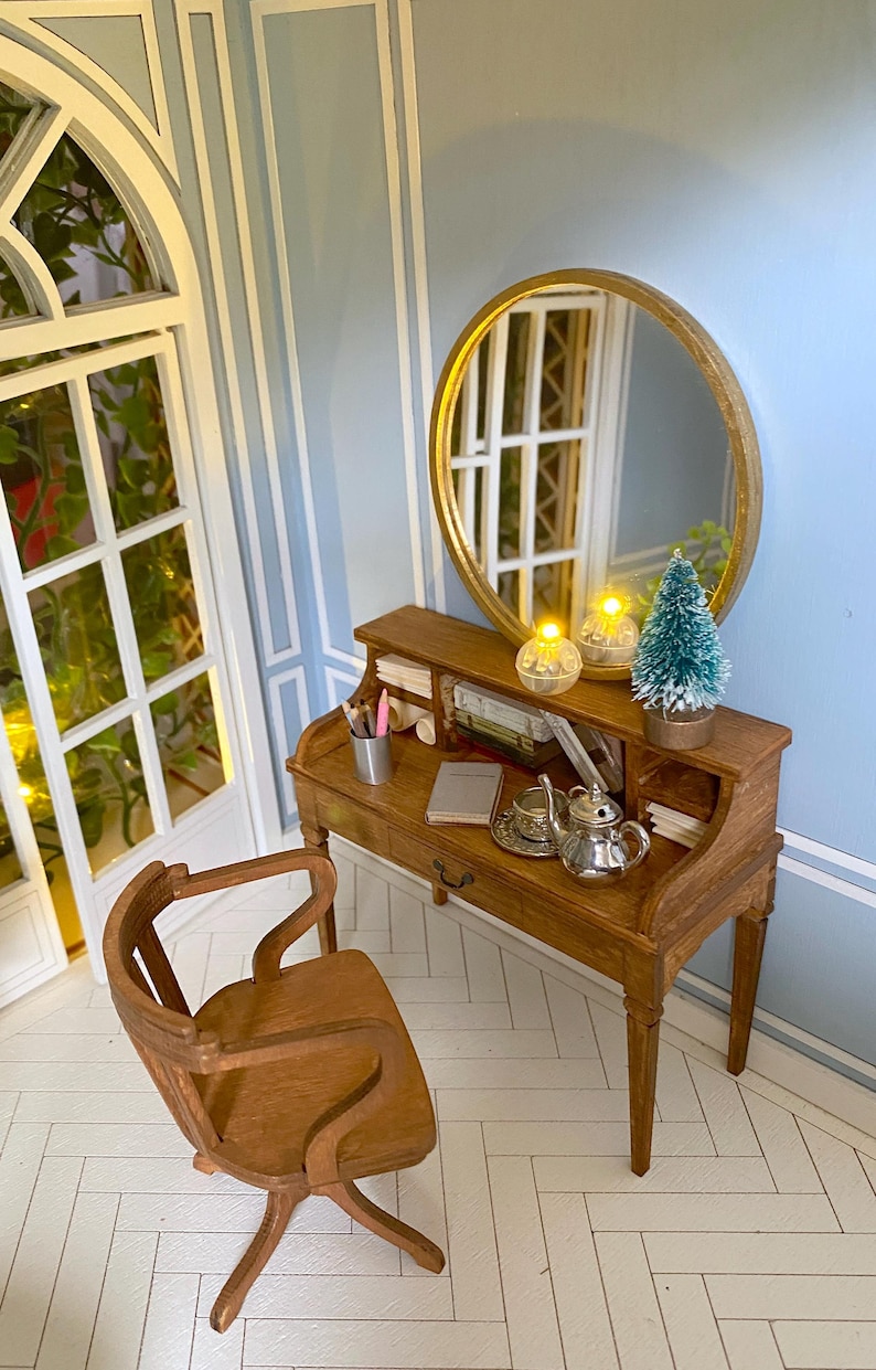 May include: Miniature wooden desk with a round mirror, chair, and accessories. The desk features a small teapot, teacup, and books. A small Christmas tree and two decorative lights sit on the desk. The chair has a curved back and legs. The room has light blue walls.