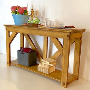 Op de afbeelding: Een houten consoletafel met een lichtbruine afwerking. De tafel heeft één plank en een X-vormige ondersteuningsstructuur. Er staan boeken, een mand en decoratieve items op de tafel.