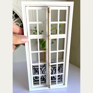 Op de afbeelding: Een miniatuur wit raam met een decoratief zwart smeedijzeren ontwerp aan de onderkant. Het raam heeft meerdere ruiten en een bamboe-achtige steun. Groene planten zijn zichtbaar door het glas en voegen een vleugje natuur toe aan de scène.