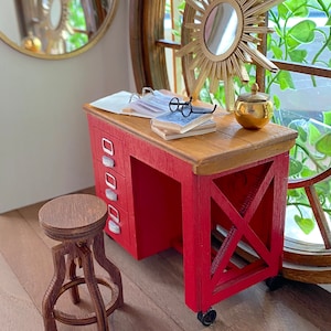 Könnte beinhalten: Miniatur-Schreibtisch in Rot mit Holzplatte, drei Schubladen und einem passenden Hocker. Ein goldfarbener Sonnenstrahlspiegel und ein runder Spiegel sind zu sehen. Ein kleines goldenes Gefäß und eine Brille liegen auf dem Schreibtisch. Die Szene spielt vor einem großen Bogenfenster.