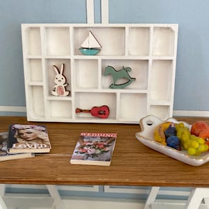 May include: A white wooden shelf with 12 compartments, featuring a variety of small wooden decorations, including a sailboat, a rocking horse, a rabbit, and a guitar. The shelf is on a wooden table with two magazines, one titled "Loved" and the other titled "Wedding". A white ceramic dish with fruit and a banana is on the table.