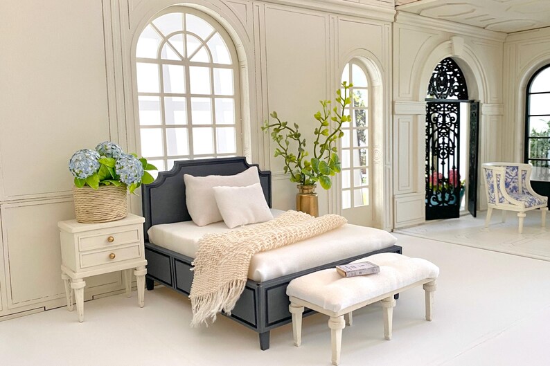 Pu&ograve; includere: Una scena di camera da letto in miniatura con un letto grigio scuro, biancheria da letto bianca e una coperta con frange color crema. Un piccolo comodino bianco contiene un cesto di ortensie blu. Una panca abbinata si trova ai piedi del letto. La stanza ha grandi finestre ad arco.