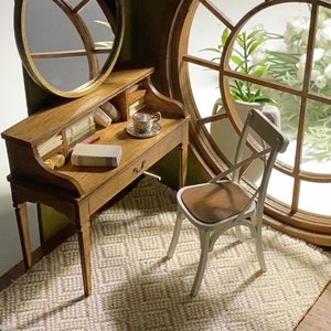 May include: Miniature wooden desk with a small drawer, books, and a teacup. A white chair with a brown seat sits on a woven rug. A round mirror and a large window with flowers complete the scene.