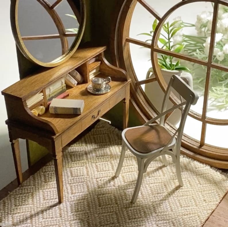 May include: Miniature wooden desk with a small drawer, books, and a teacup. A white chair with a brown seat sits on a woven rug. A round mirror and a large window with flowers complete the scene.
