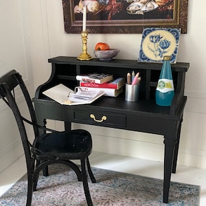 May include: Miniature black desk with a matching chair, set against a white wall. The desk holds books, pencils, and a bottle of water. A framed still life painting and a decorative tile are on the wall above the desk. A patterned rug is on the floor.