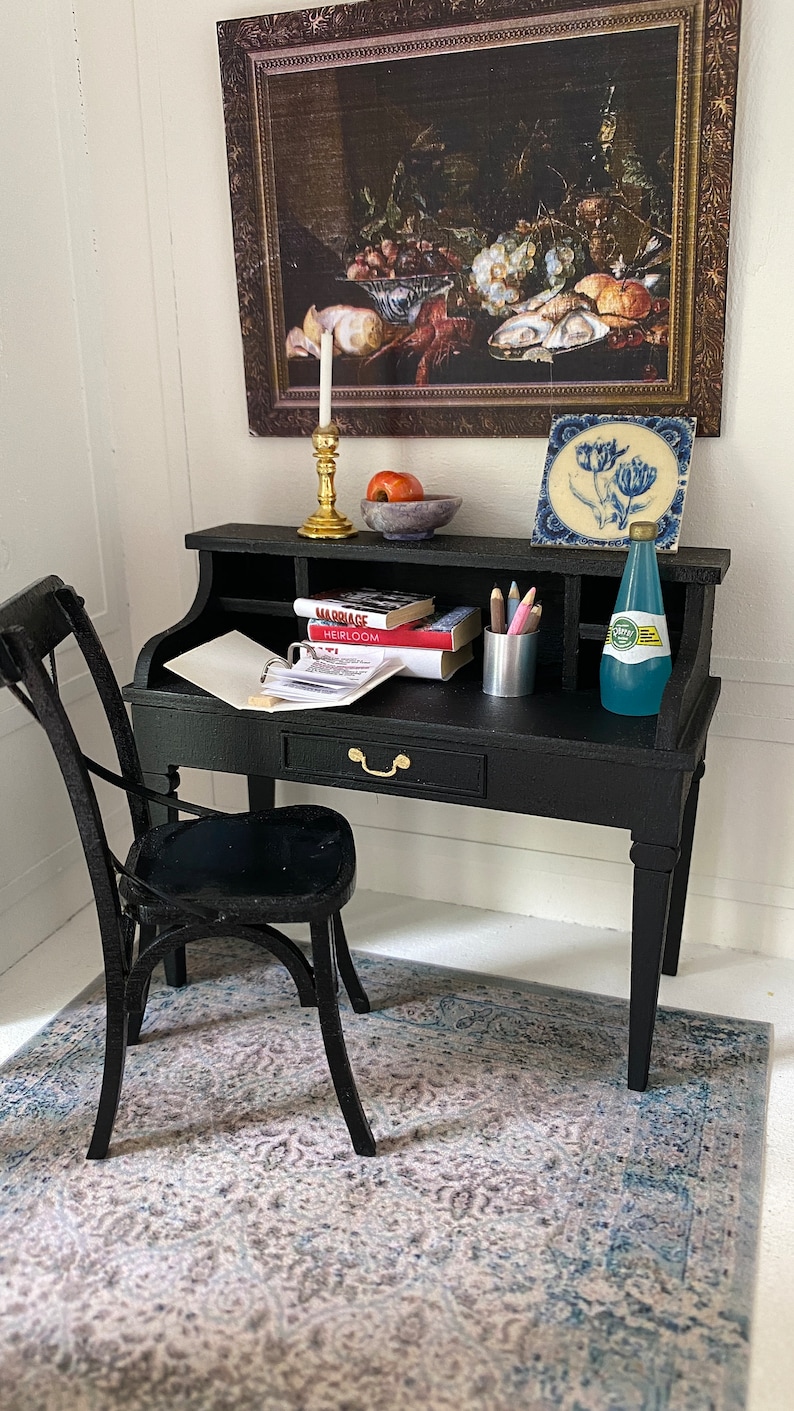 May include: Miniature black desk with a matching chair, set against a white wall. The desk holds books, pencils, and a bottle of water. A framed still life painting and a decorative tile are on the wall above the desk. A patterned rug is on the floor.