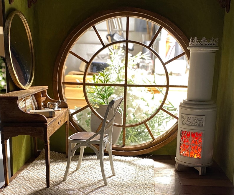 May include: A miniature room scene featuring a wooden desk with a mirror, a white chair, and a decorative white stove. A large round window offers a view of the outside. The room has a cozy, inviting atmosphere.