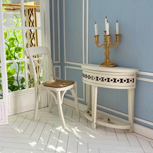 May include: A white wooden chair with a brown seat sits next to a white half-round table with a gold candle holder with white candles. The table has a decorative design around the edge.