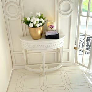 May include: White half-round table with carved details, a gold planter with white flowers, a candle with a floral design, and two books on top. The table is in a white room with white tiled flooring.