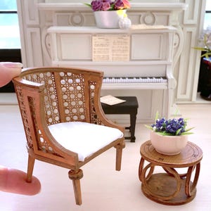 Peut inclure: Une chaise miniature en bois avec un coussin blanc et un dossier en osier. La chaise est posée sur une surface blanche à côté d'une table miniature en bois avec un plateau rond et une base en osier. Un petit pot blanc avec des fleurs violettes est posé sur la table.