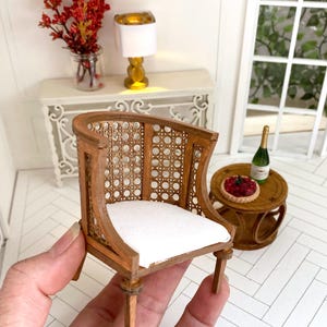 K&ouml;nnte beinhalten: Ein Miniatur-Holzstuhl mit einem wei&szlig;en Kissen und einer geflochtenen R&uuml;ckenlehne aus Rohrgeflecht. Der Stuhl ist braun und hat eine gebogene R&uuml;ckenlehne.