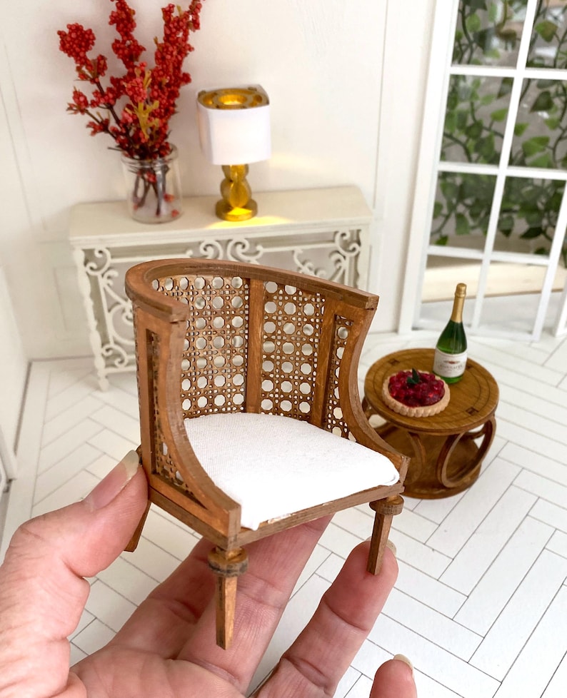 K&ouml;nnte beinhalten: Ein Miniatur-Holzstuhl mit einem wei&szlig;en Kissen und einer geflochtenen R&uuml;ckenlehne aus Rohrgeflecht. Der Stuhl ist braun und hat eine gebogene R&uuml;ckenlehne.