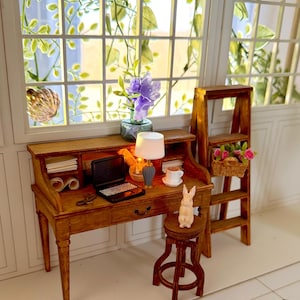 May include: Miniature wooden desk with a laptop, lamp, and coffee cup. A small wooden stool with a bunny figurine sits in front of the desk. A small wooden ladder with a basket of flowers is to the right. A window with greenery is in the background.
