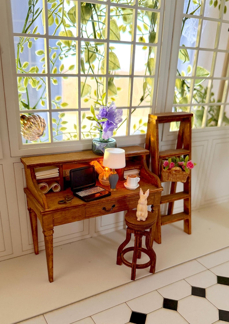 May include: Miniature wooden desk with a laptop, lamp, and coffee cup. A small wooden stool with a bunny figurine sits in front of the desk. A small wooden ladder with a basket of flowers is to the right. A window with greenery is in the background.