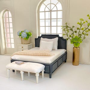 Pu&ograve; includere: Scena di camera da letto in miniatura con un letto nero, biancheria da letto bianca e una coperta color crema. Una panca bianca &egrave; ai piedi del letto. Un comodino con fiori blu e un vaso dorato con verde completano la scena.
