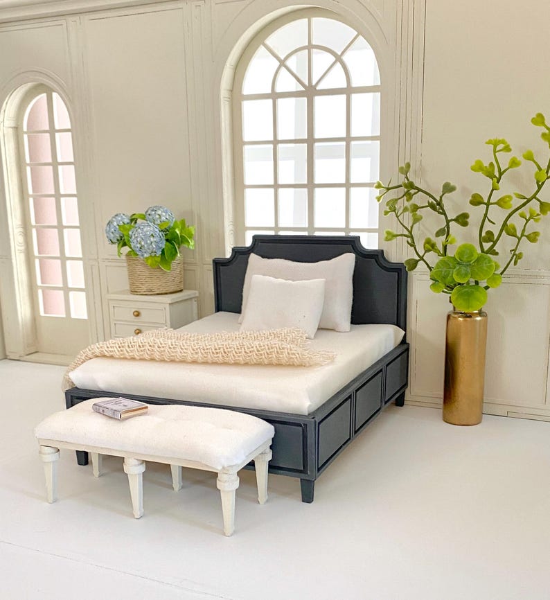 Pu&ograve; includere: Scena di camera da letto in miniatura con un letto nero, biancheria da letto bianca e una coperta color crema. Una panca bianca &egrave; ai piedi del letto. Un comodino con fiori blu e un vaso dorato con verde completano la scena.