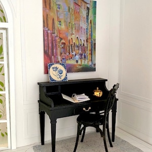 May include: Miniature black desk and chair set with a colorful street scene painting and a blue and white tile. A small gold-colored container sits on the desk, along with papers. The desk has a drawer and a curved back.