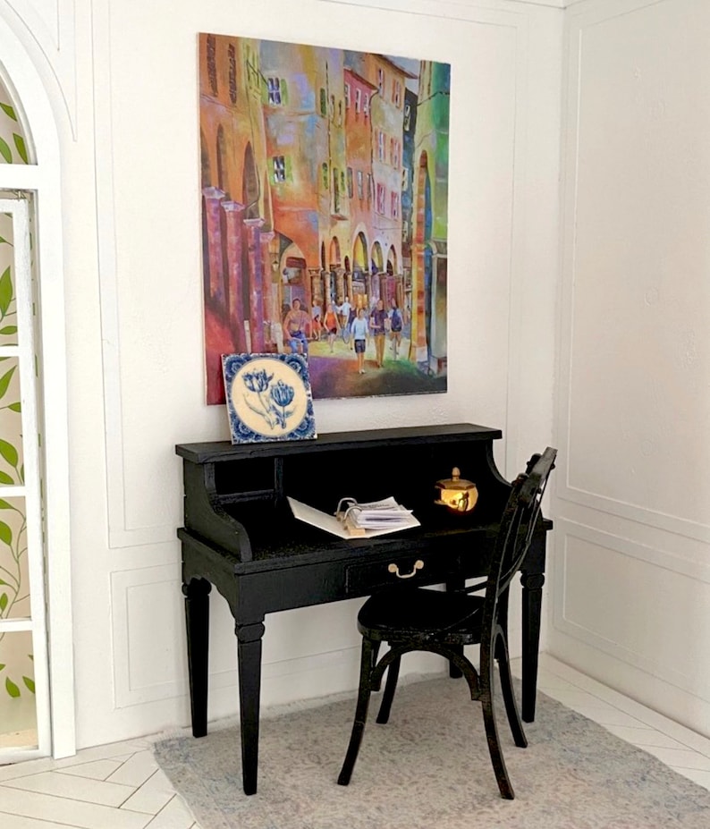 May include: Miniature black desk and chair set with a colorful street scene painting and a blue and white tile. A small gold-colored container sits on the desk, along with papers. The desk has a drawer and a curved back.