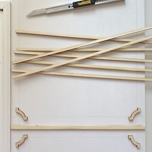 Pu&ograve; includere: Una superficie bianca con un disegno a matita di una cornice. Ci sono strisce di legno e un coltello multiuso sulla superficie. Il coltello multiuso ha la scritta "OLFA" su di esso.