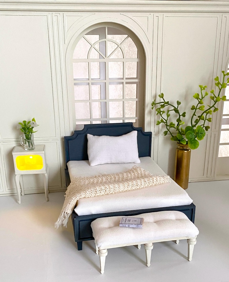 Pu&ograve; includere: Set da camera da letto in miniatura con un letto blu navy, biancheria da letto bianca e una coperta color crema. Una panca bianca si trova ai piedi del letto e un piccolo comodino con luce gialla &egrave; a sinistra. Un vaso con verde &egrave; a destra.