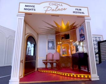 Pode incluir: Um modelo em miniatura de um cinema com um tapete vermelho, uma bilheteria e duas filas de assentos. A placa acima da entrada diz "Cinema Palace" com "Movie Nights" à esquerda e "Film Festival" à direita.