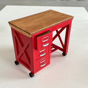 Könnte beinhalten: Ein kleiner roter Holzschreibtisch mit einer hellbraunen Tischplatte. Der Schreibtisch hat drei Schubladen mit silbernen Griffen und steht auf kleinen schwarzen Rädern. Der Schreibtisch hat eine X-förmige Stütze an den Seiten.