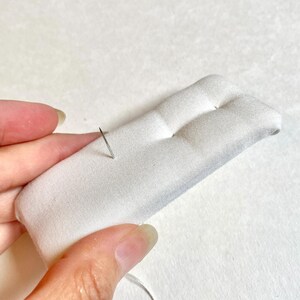 Pu&ograve; includere: Un portaspilli rettangolare bianco con due sezioni arrotondate. Un ago d'argento &egrave; inserito nel tessuto, con filo attaccato. Il cuscino &egrave; tenuto da una mano. Il materiale &egrave; morbido e trapuntato.