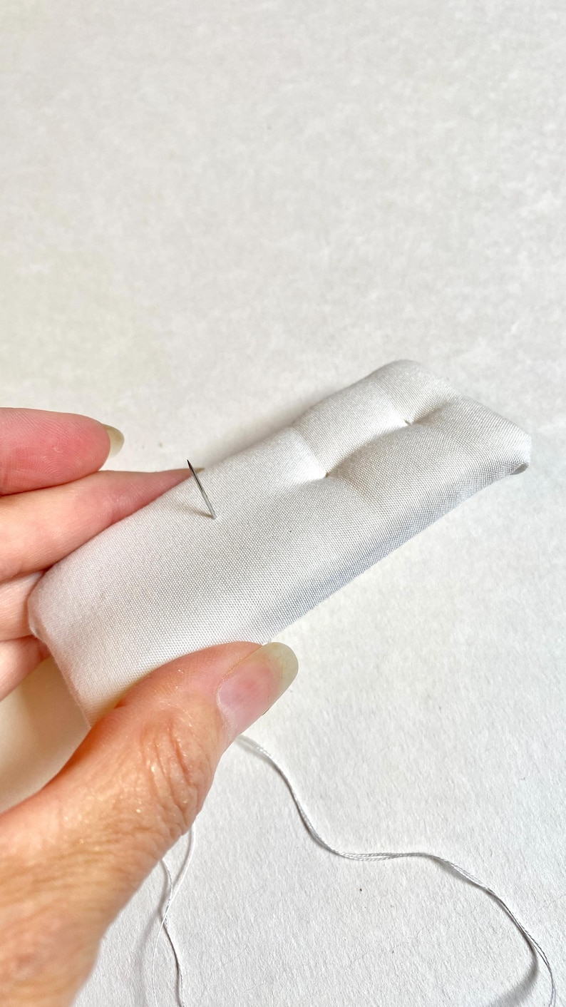 Pu&ograve; includere: Un portaspilli rettangolare bianco con due sezioni arrotondate. Un ago d'argento &egrave; inserito nel tessuto, con filo attaccato. Il cuscino &egrave; tenuto da una mano. Il materiale &egrave; morbido e trapuntato.