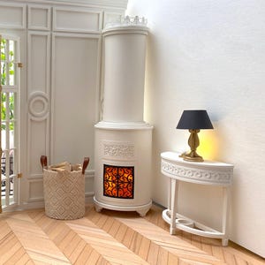 Op de afbeelding: Een witte miniatuur open haard met een decoratieve bovenkant en een gloeiende vuurhaard. Een kleine tafel met een zwarte lamp en een geweven mand met brandhout staan in de buurt. De kamer heeft een parketvloer en witte muren.