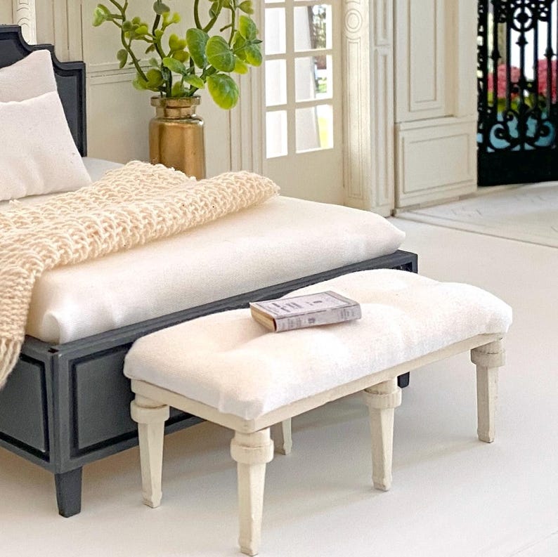 Pu&ograve; includere: Scena di camera da letto in miniatura con un letto con struttura grigio scuro, biancheria da letto bianca e una coperta lavorata a maglia color crema. Una piccola panca imbottita con un libro poggia ai piedi del letto. Un vaso dorato con vegetazione &egrave; visibile.