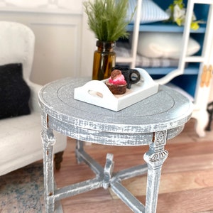 May include: A white tray with a small dessert and a cup of coffee sits on a gray distressed round side table with carved legs.