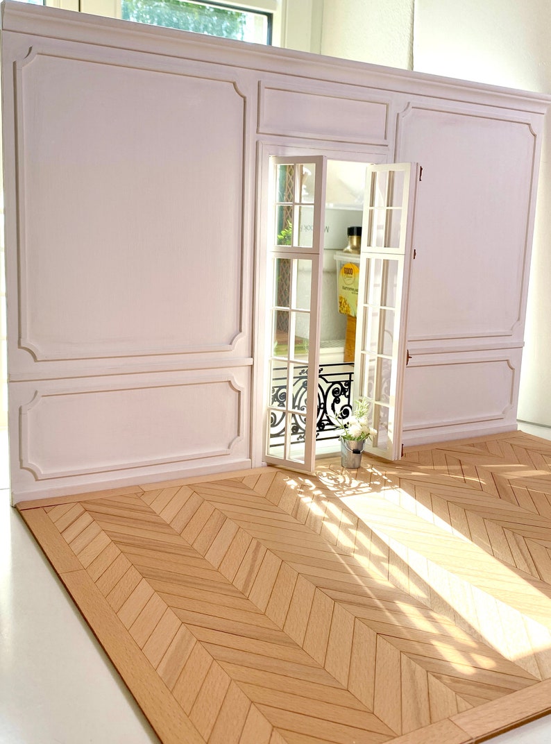 Pu&ograve; includere: Una stanza di una casa delle bambole bianca con una parete a boiserie bianca e una porta con pannelli di vetro che conduce a un balcone. Il pavimento &egrave; fatto di assi di legno marrone chiaro disposte a spina di pesce.
