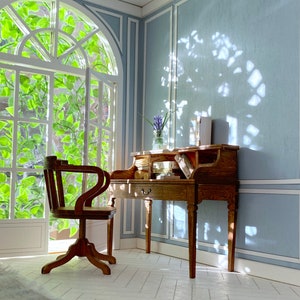 May include: A miniature wooden desk and chair set in a room with a large window. The desk has drawers and compartments, and the chair has a curved back. The room has light blue walls and white trim, with sunlight streaming through the window.