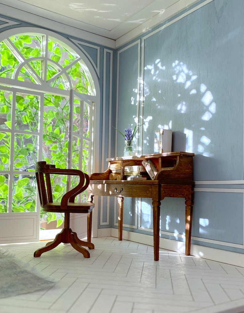May include: A miniature wooden desk and chair set in a room with a large window. The desk has drawers and compartments, and the chair has a curved back. The room has light blue walls and white trim, with sunlight streaming through the window.