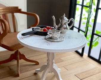 May include: A white, round table with a decorative design on the top. A silver teapot, cup, and saucer are on the table. The table has three legs and is made of wood.