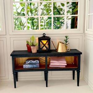 May include: A miniature black table with gold detailing, featuring a small lantern, a gold watering can, and potted plants. The lower shelves display books and pink linens. The scene is set against a window with greenery.