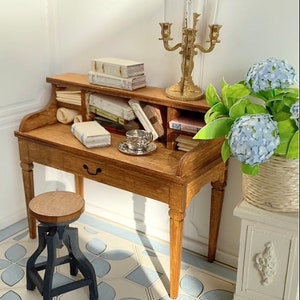 May include: A miniature wooden desk with a drawer, holding books, a teacup, and a gold candelabra. A small stool sits in front of the desk. A vase of blue flowers is to the right. The floor has a blue and white tile pattern.