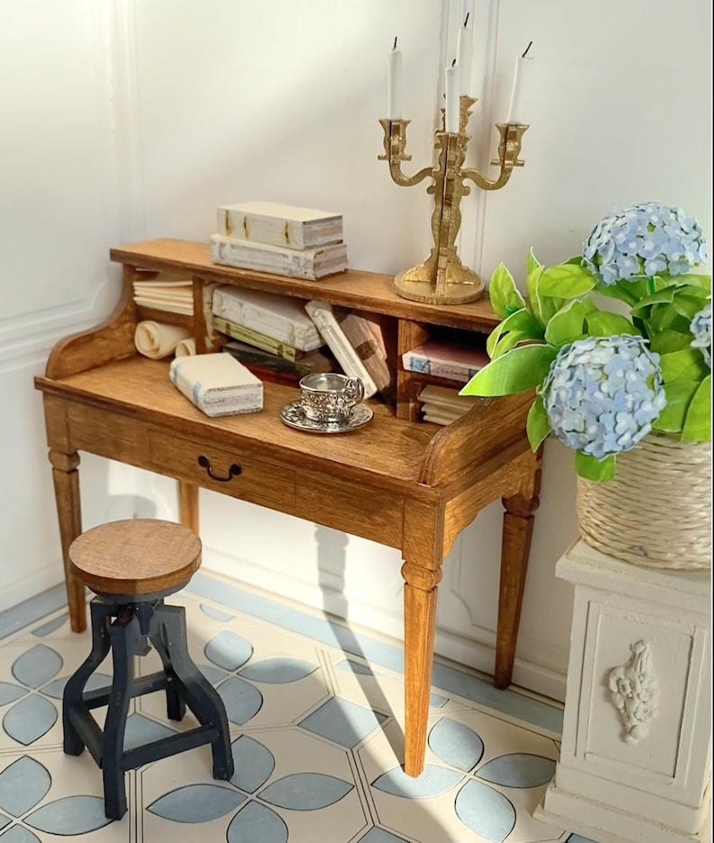 May include: A miniature wooden desk with a drawer, holding books, a teacup, and a gold candelabra. A small stool sits in front of the desk. A vase of blue flowers is to the right. The floor has a blue and white tile pattern.