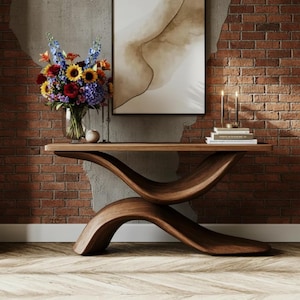 Op de afbeelding: Een moderne console van donker hout met een uniek, gebogen ontwerp. De tafel toont een vaas met kleurrijke bloemen, kaarsen en boeken. De achtergrond is een bakstenen muur en abstracte kunst. De tafel is ongeveer 150 cm lang.