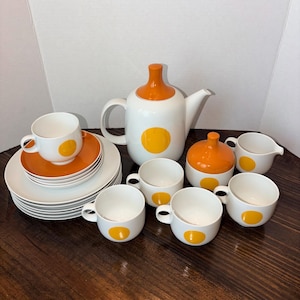 May include: A white and orange ceramic tea set with a teapot, cups, saucers, plates, a sugar bowl, and a creamer. The set features a large yellow dot on each piece. The teapot has a rounded shape with an orange lid.