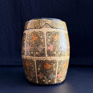 May include: A decorative, barrel-shaped container with a lid. The container is black with intricate gold floral patterns and accents in orange, pink, and blue. The design features stylized leaves and vines, creating a vintage aesthetic.