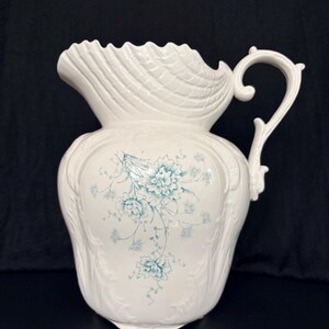 May include: A white ceramic pitcher with a ruffled rim and ornate handle. The pitcher features a floral design in shades of teal. The body of the pitcher has a bulbous shape with embossed detailing.