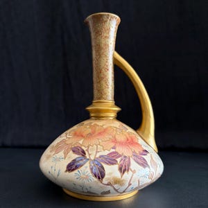 May include: A decorative ceramic pitcher with a bulbous body and a slender, curved handle. The pitcher features a cream-colored base adorned with floral designs in shades of pink, purple, and gold. The handle and spout are gold and brown.