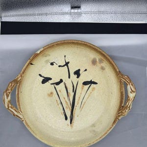 May include: A round ceramic serving dish with two textured, branch-like handles on opposite sides. The dish features a speckled cream-colored glaze with a hand-painted black abstract floral design in the center and subtle brown accents.