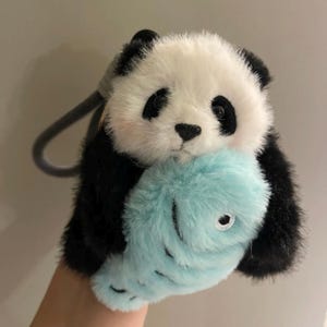 May include: A soft toy panda bear with black and white fur, clutching a light blue stuffed fish. The panda has black eyes and ears, with a grey loop attached to its head. The fish has a white embroidered eye.