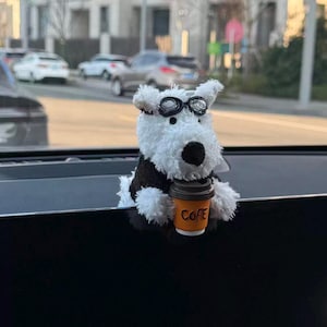 May include: A fluffy white dog ornament wearing black spectacles and a black waistcoat, holding a small coffee cup with the word "COFE" printed on it. The dog is a car accessory, designed to sit on a dashboard.