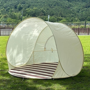 May include: A beige pop-up tent with a brown and white striped floor is set up on a grassy lawn. The tent has a rounded shape and is open at the front, revealing the interior. The background features trees and a fence.