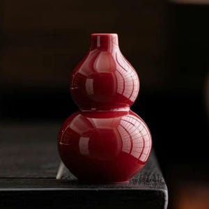 Puede incluir: Un jarrón brillante en forma de calabaza roja con un diseño de doble bulbo. El jarrón tiene una superficie lisa y reflectante, y el fondo oscuro realza su color vibrante. El jarrón es un objeto decorativo.