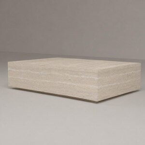 May include: A rectangular coffee table crafted from beige travertine stone. The table features a low-profile, block-like design with a smooth, flat top surface and visible natural horizontal veining patterns throughout the porous stone material.