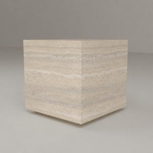 May include: A cube-shaped pedestal crafted from beige travertine stone. The surface displays natural horizontal banding and a porous, speckled texture characteristic of the material, forming a solid geometric block with sharp, clean edges.